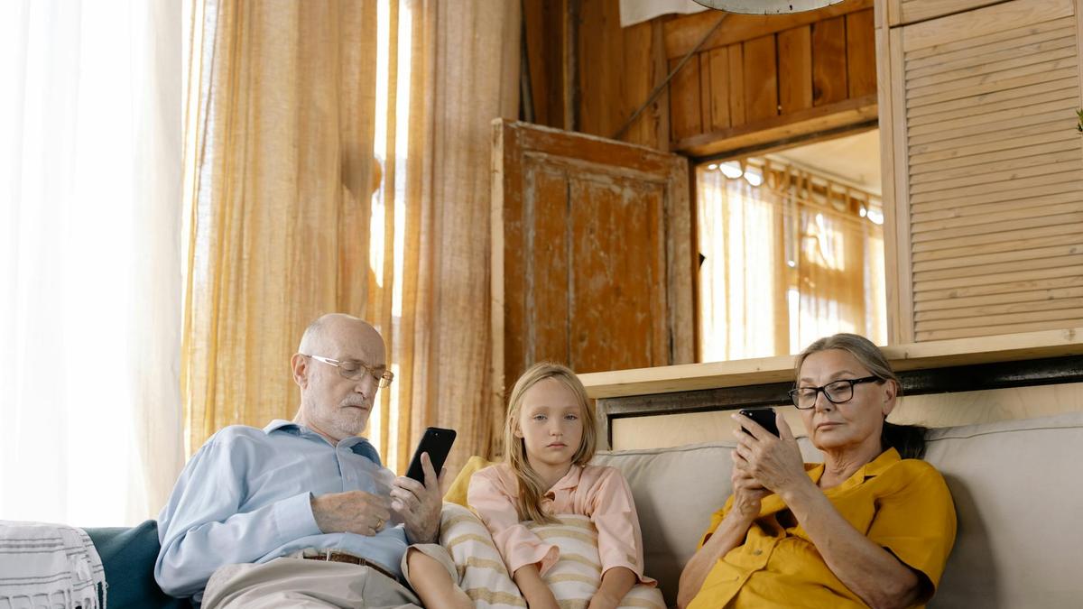 A young girl and her grandparents sitting on a sofa, using smartphones in a cozy living room.