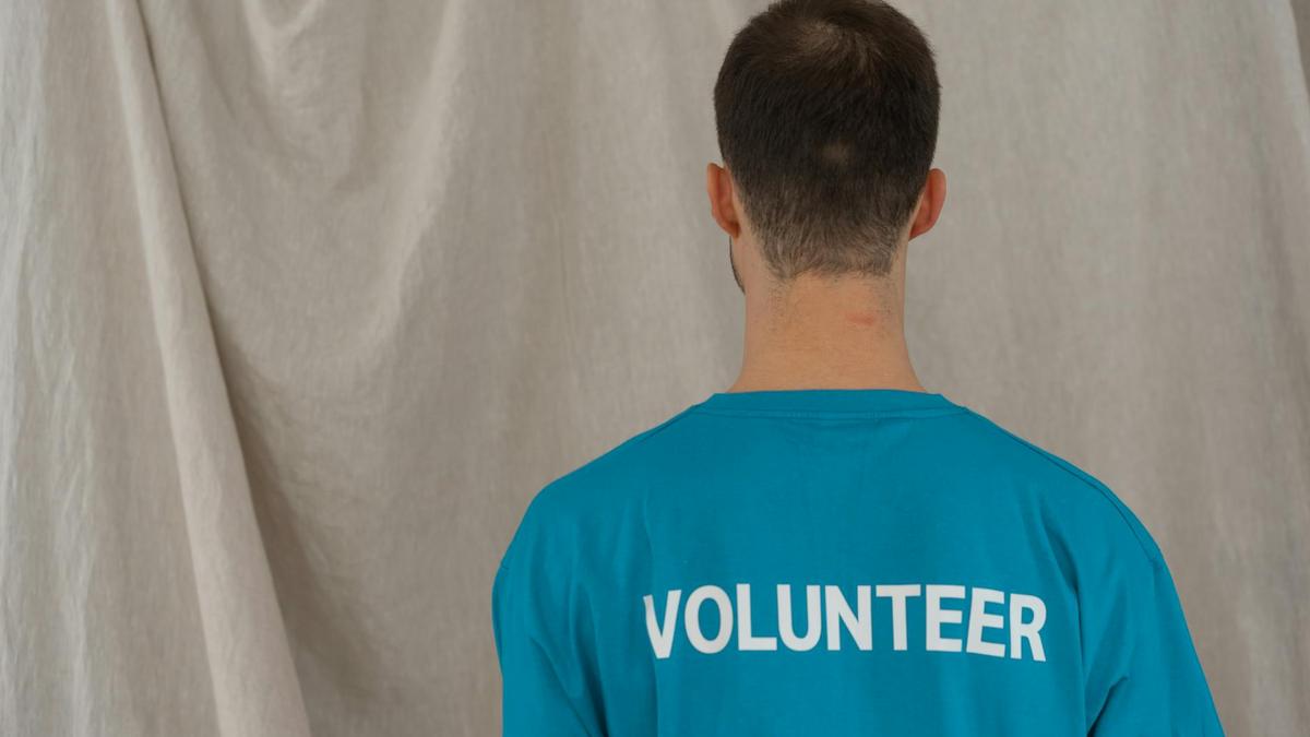 A man in a blue volunteer shirt seen from behind, indoors.