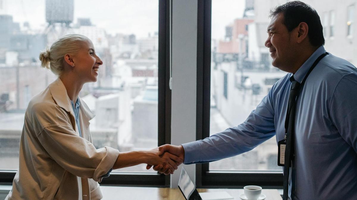 Side view of diverse coworkers in formal clothes standing near table with cup of coffee and laptop and shaking hands after successful business project in office with glass wall