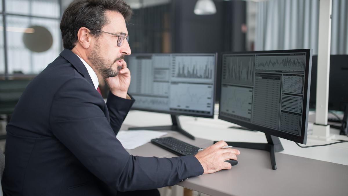 Businessman analyzing stock market data on dual monitors in a modern office setting.