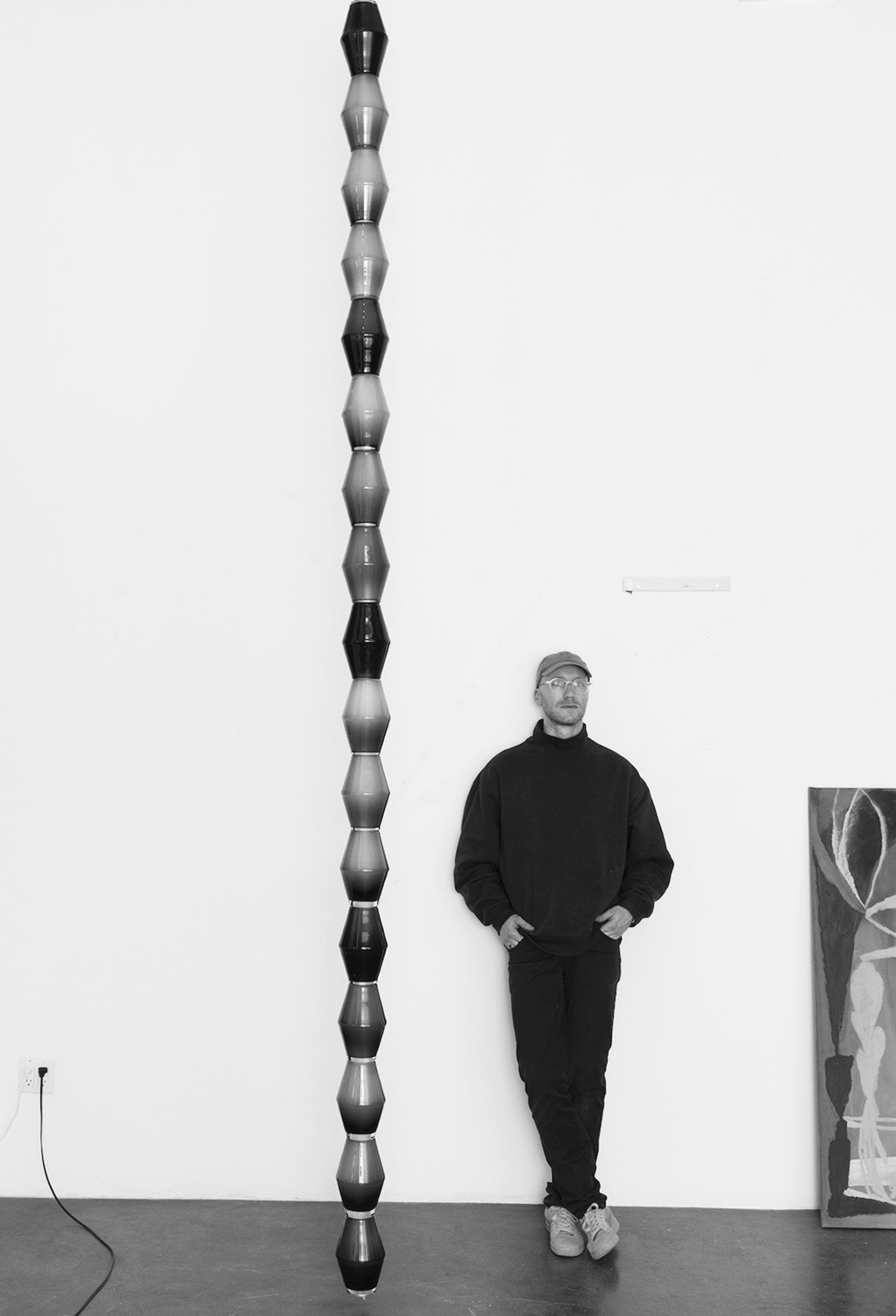 Tron Meyer standing against a white wall next to a tall stacked sculpture