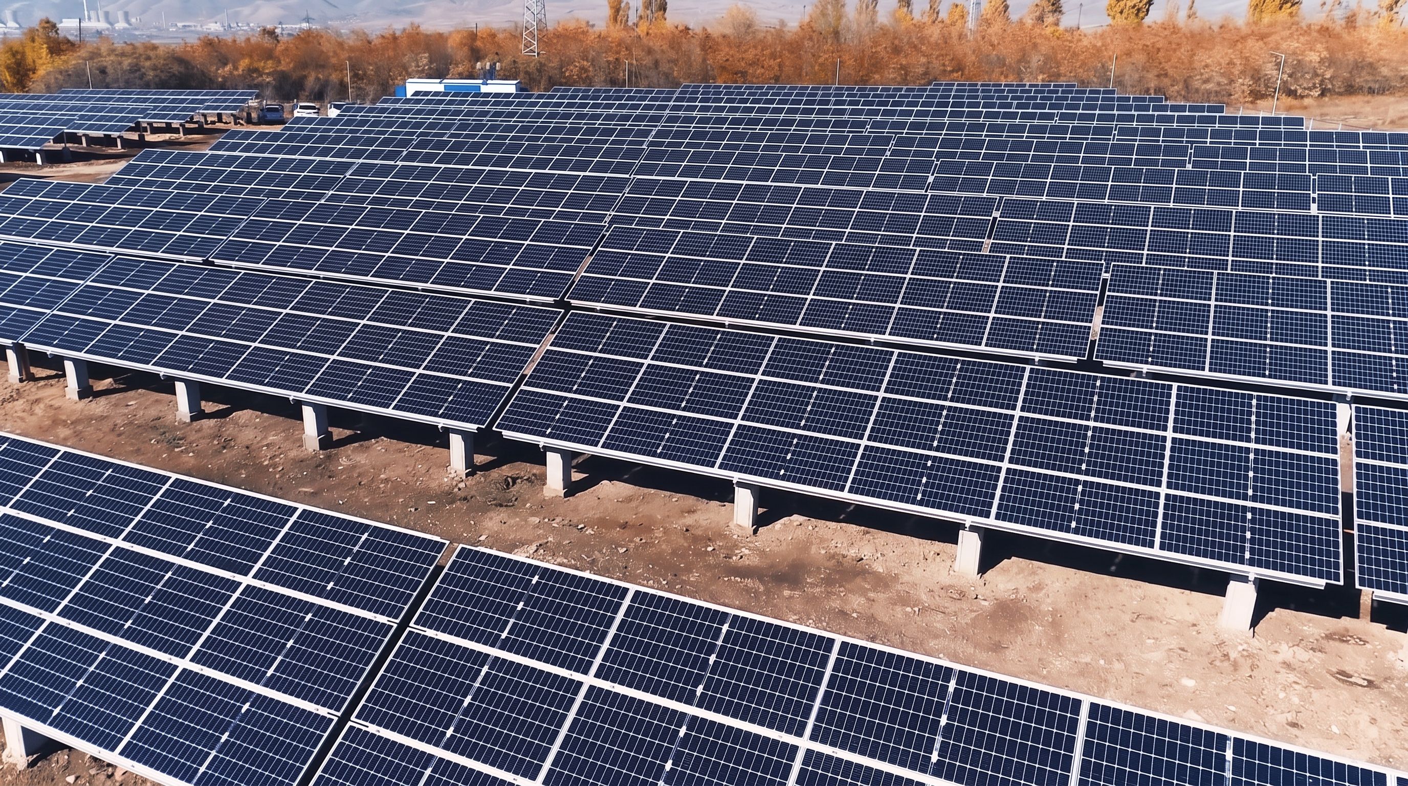 Rows of solar panels on concrete footings at the ARSUN power plant in Hrazdan, with bare autumn trees and hills behind.