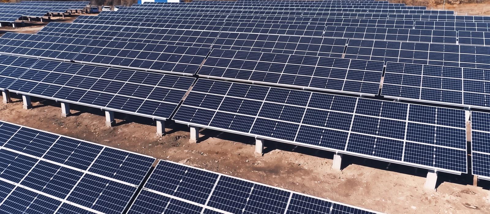 Rows of solar panels on concrete footings at the ARSUN power plant in Hrazdan, with bare autumn trees and hills behind.