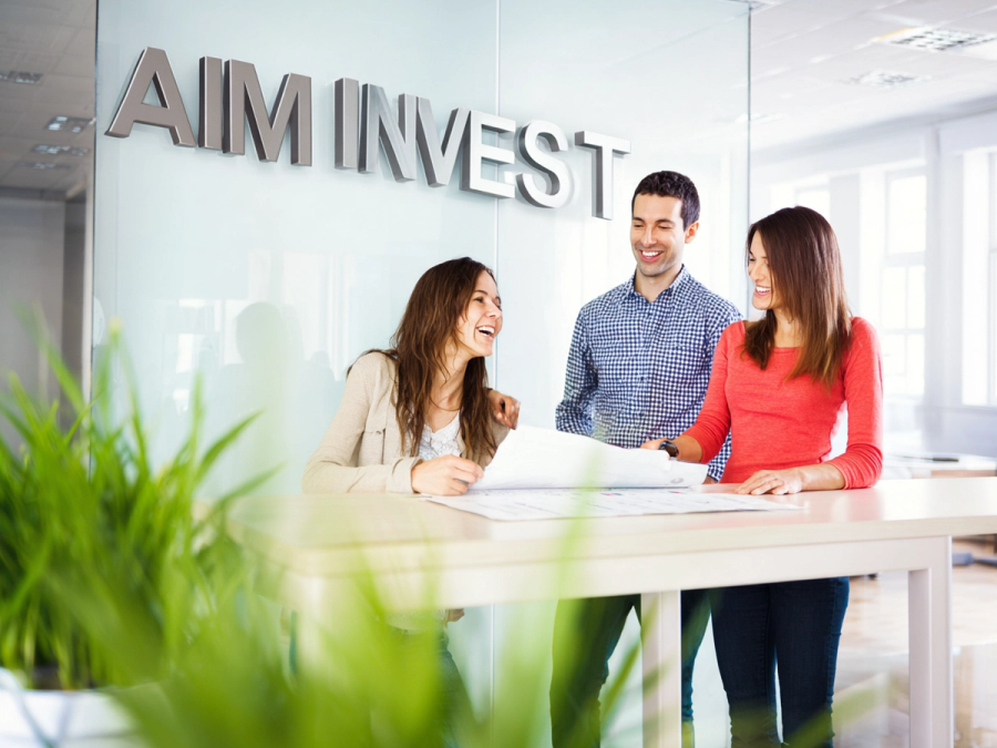 Three colleagues talking over documents in an office beneath an AIM Invest sign.