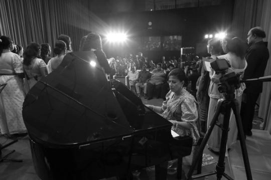 Darshani Chandrasena plays a grand piano on stage, singers with song sheets behind her