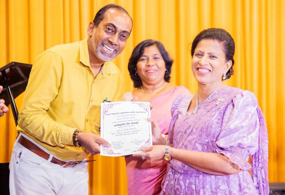 Smiling, Darshani Chandrasena holds up a certificate with a guest before golden curtains