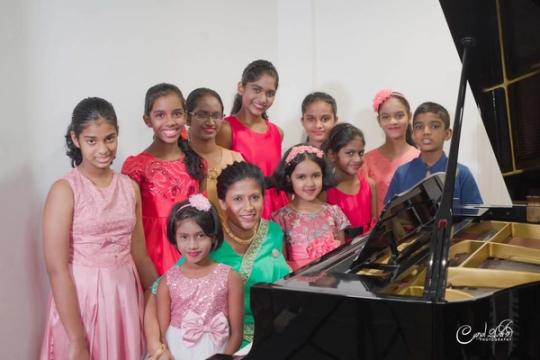 Ten children in party clothes crowd around Darshani Chandrasena at the grand piano
