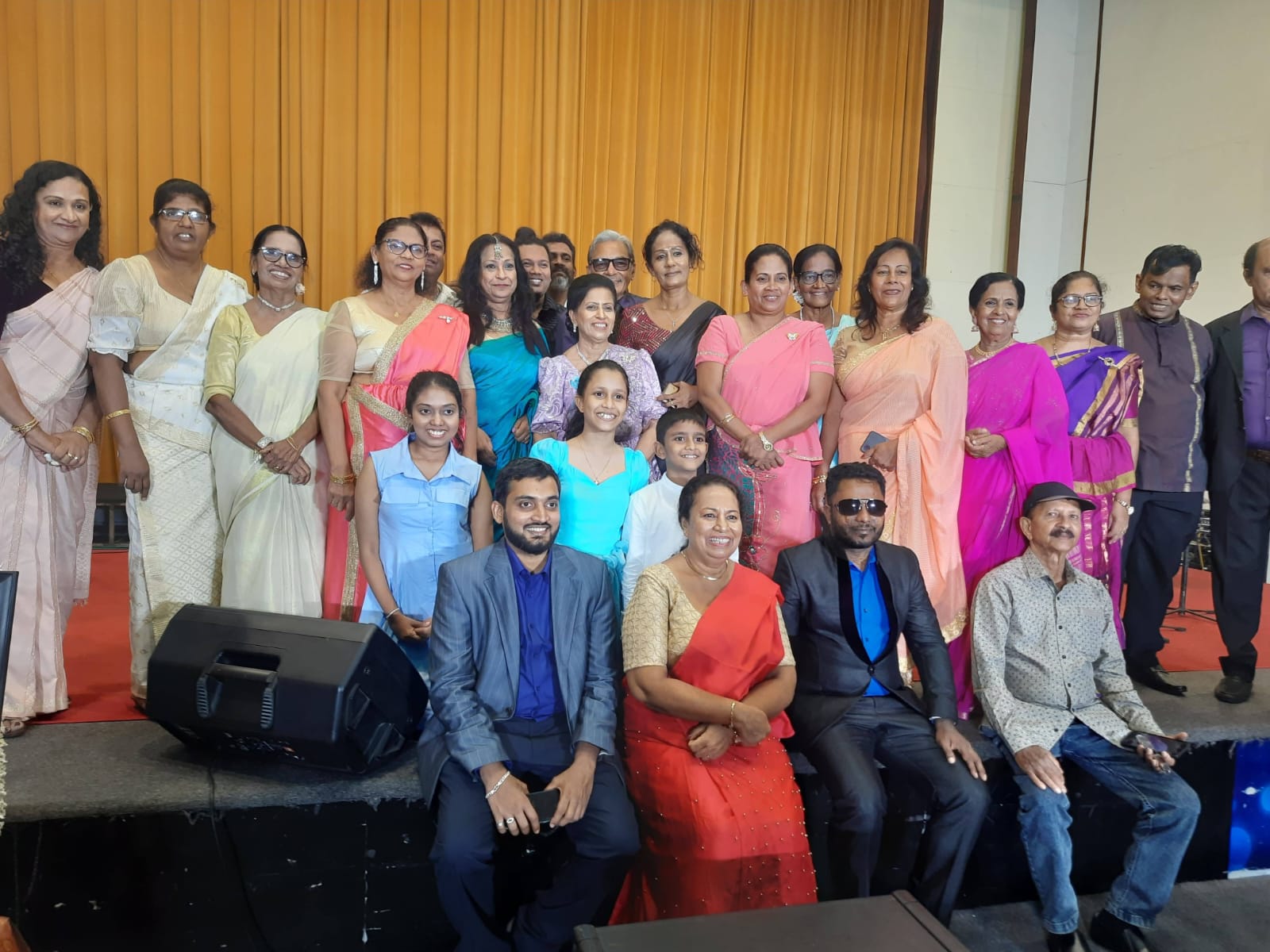 About thirty people, many in sarees, grouped on stage before a gold curtain