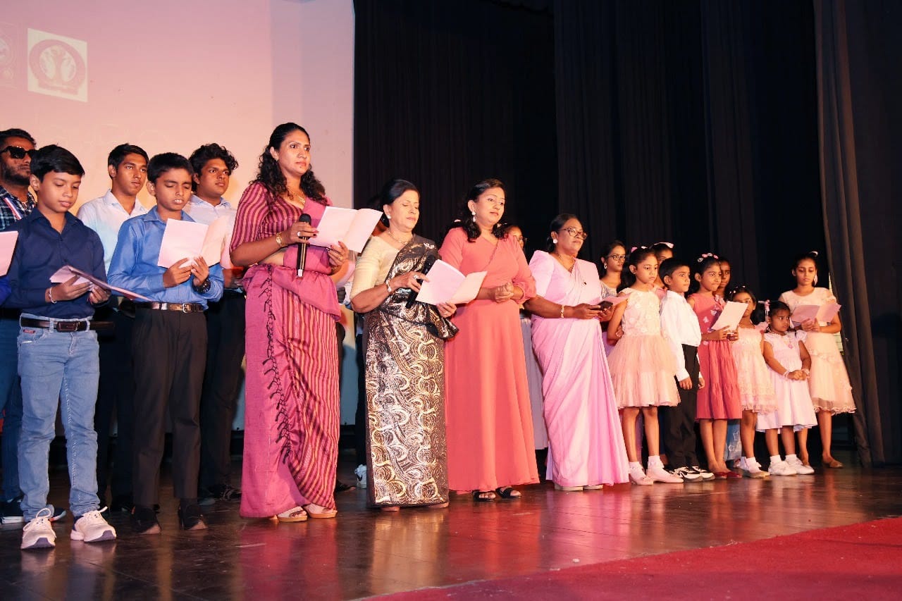 Children and adults in a line on stage, singing from song sheets, one at a microphone