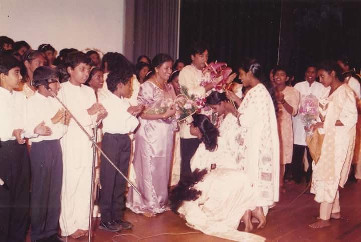 A young woman kneels before a woman in white on stage; students nearby hold bouquets