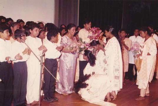 A young woman kneels before a woman in white on stage; students nearby hold bouquets
