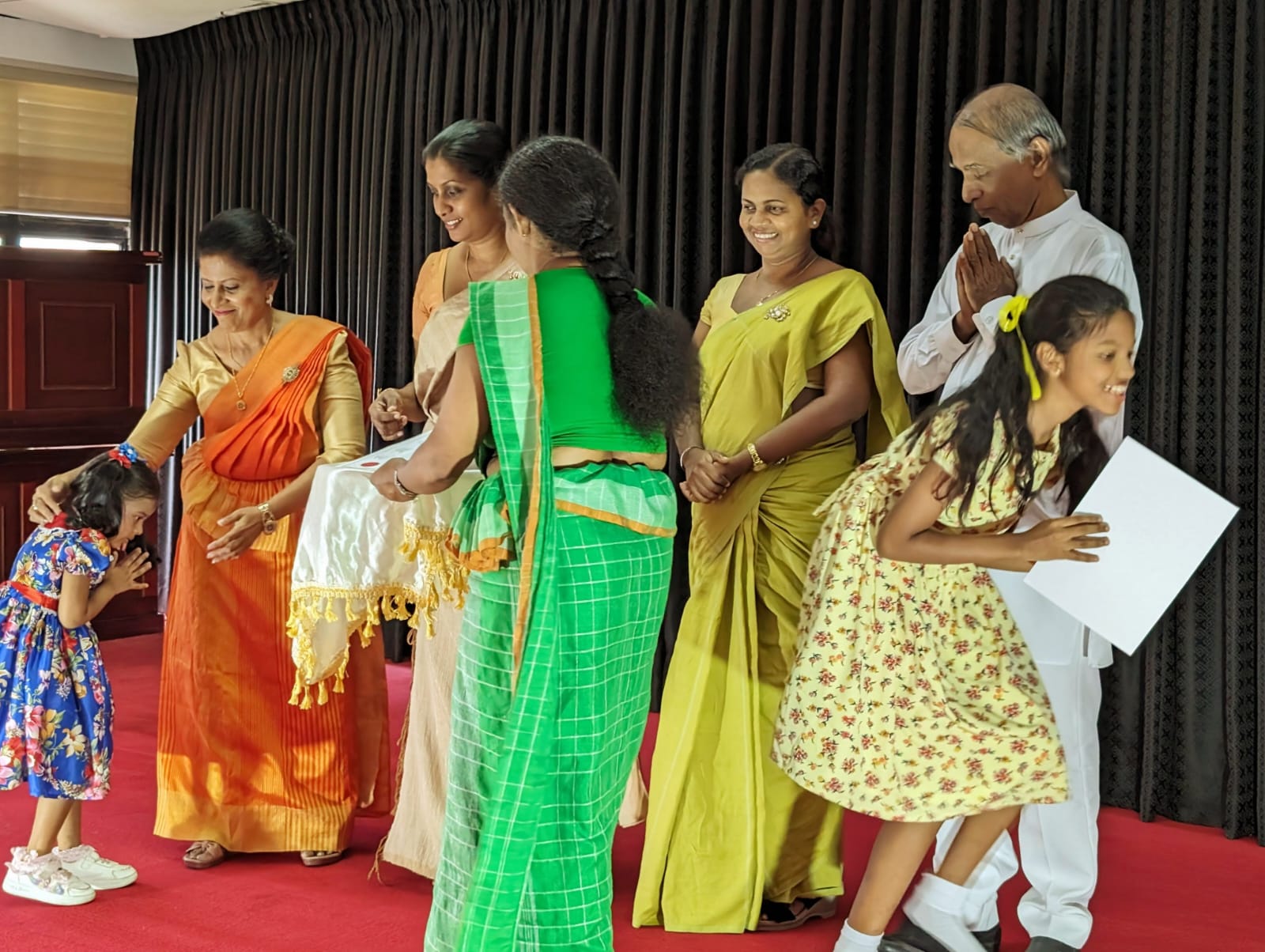 A small girl bows on stage as a woman in an orange sari touches her head