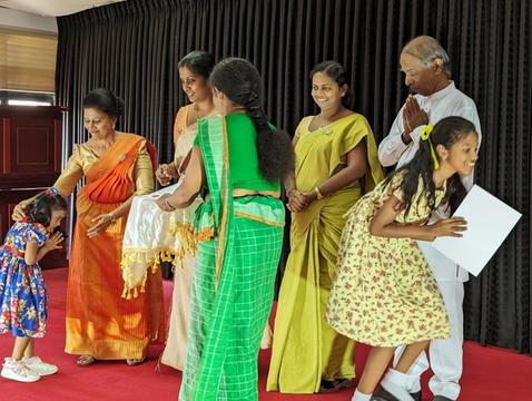 A small girl bows on stage as a woman in an orange sari touches her head