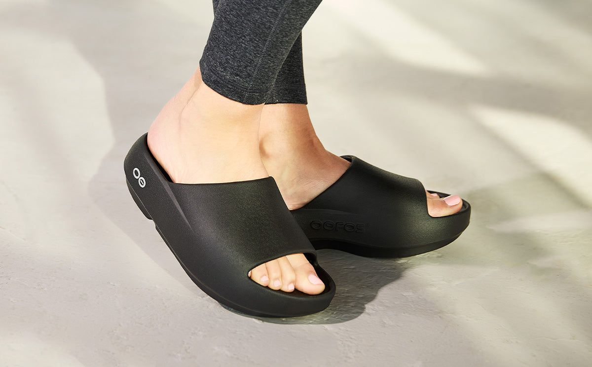 Black OOFOS slide sandals worn on a light stone floor.