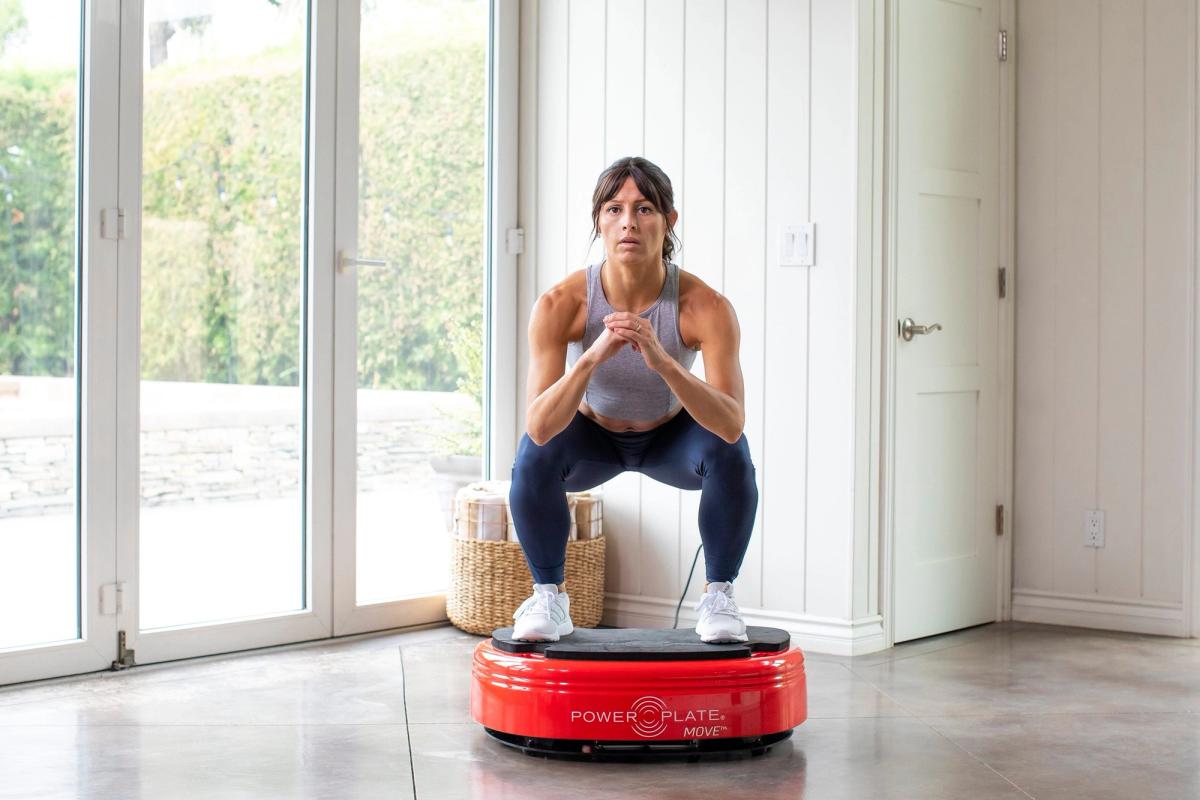A woman squats on a red vibration plate in a bright living room.