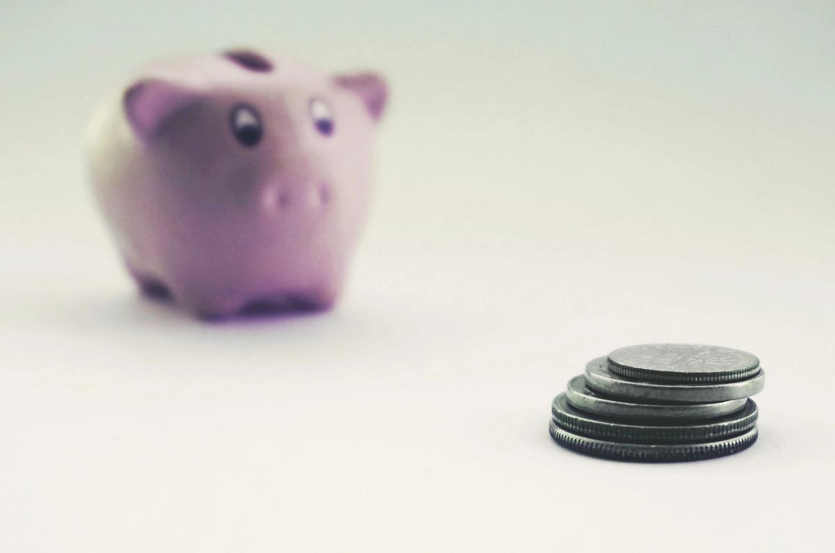 A stack of coins in focus in front of a pink piggy bank on a white surface