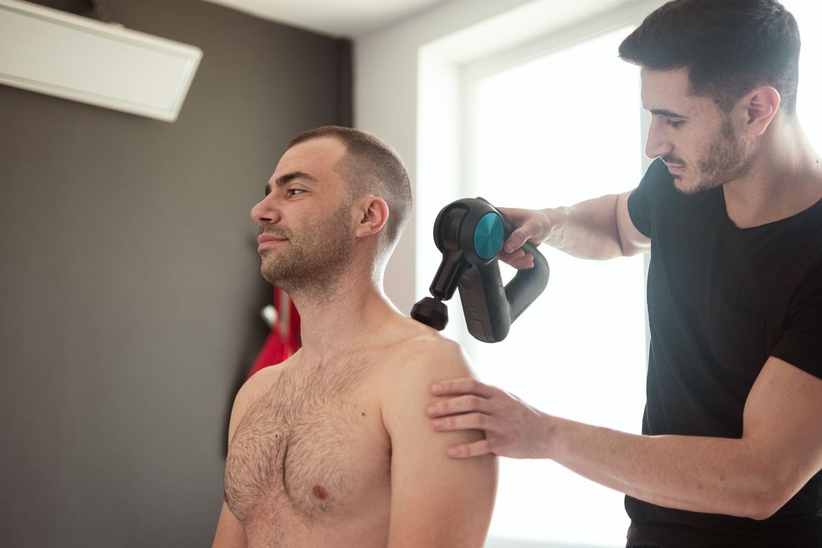 Physiotherapist using a massage gun on a seated man's shoulder near his neck.