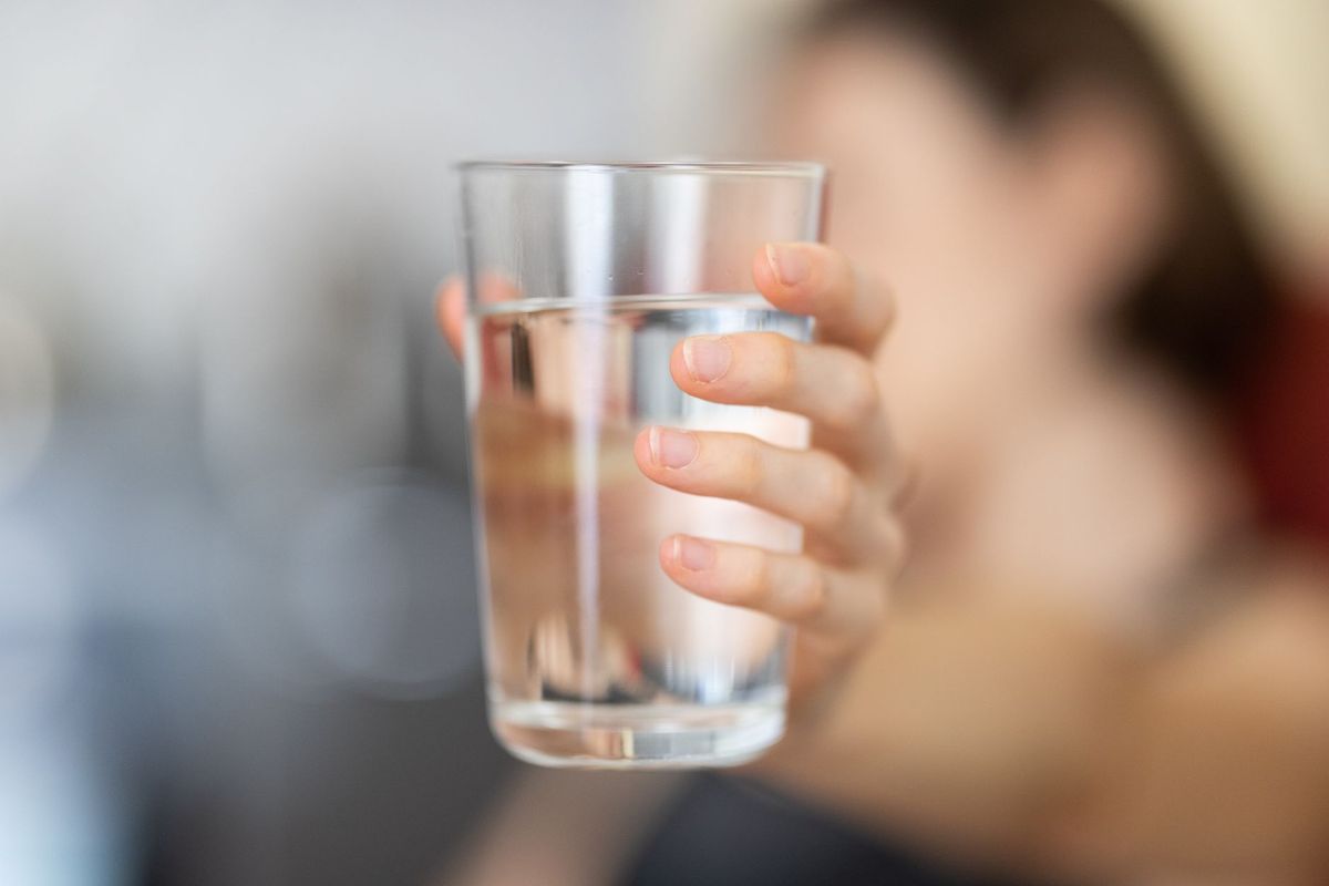A person holding a clear glass of water at eye level indoors, with a softly blurred background.