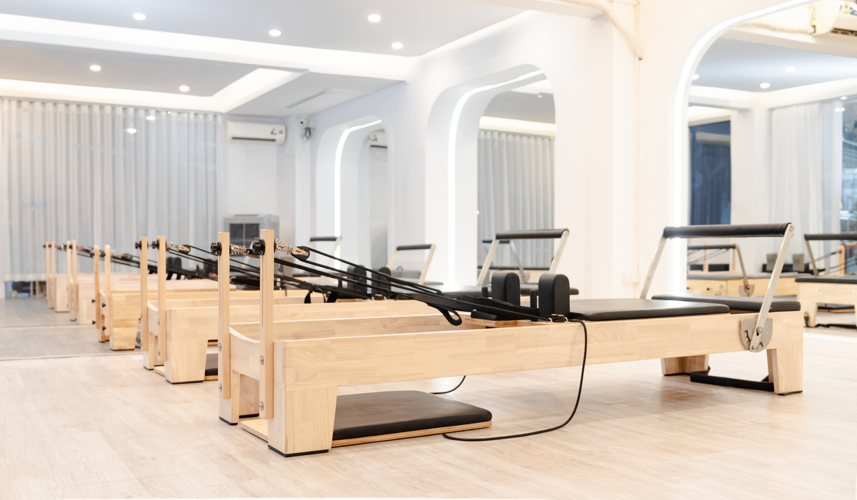 A row of wooden Pilates reformers with black carriages, footbars and straps in a bright studio.