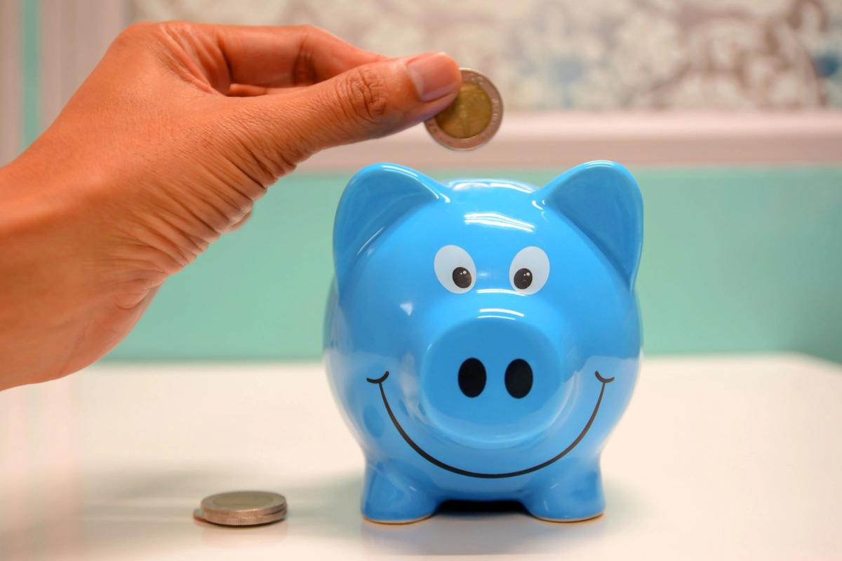 A hand dropping a coin into a smiling blue piggy bank on a white table