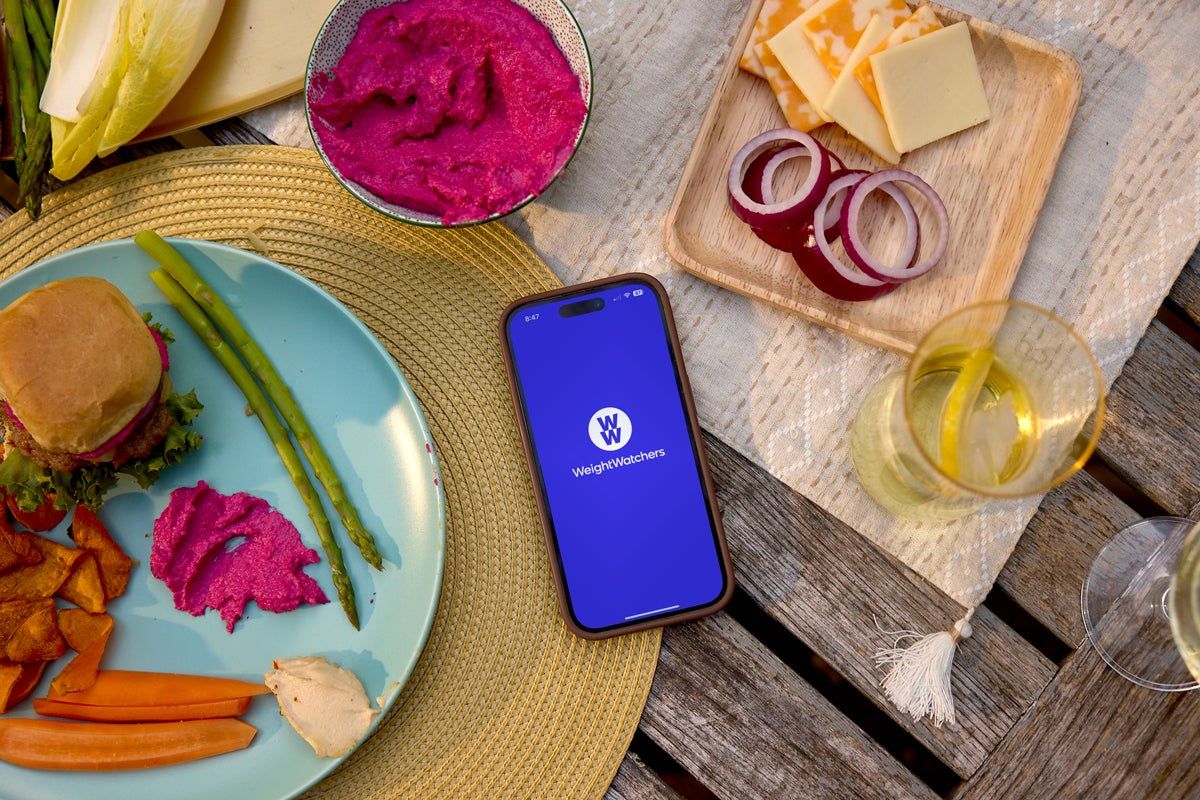 A phone showing the WeightWatchers logo on an outdoor table set with a burger, vegetables, dips and cheese.