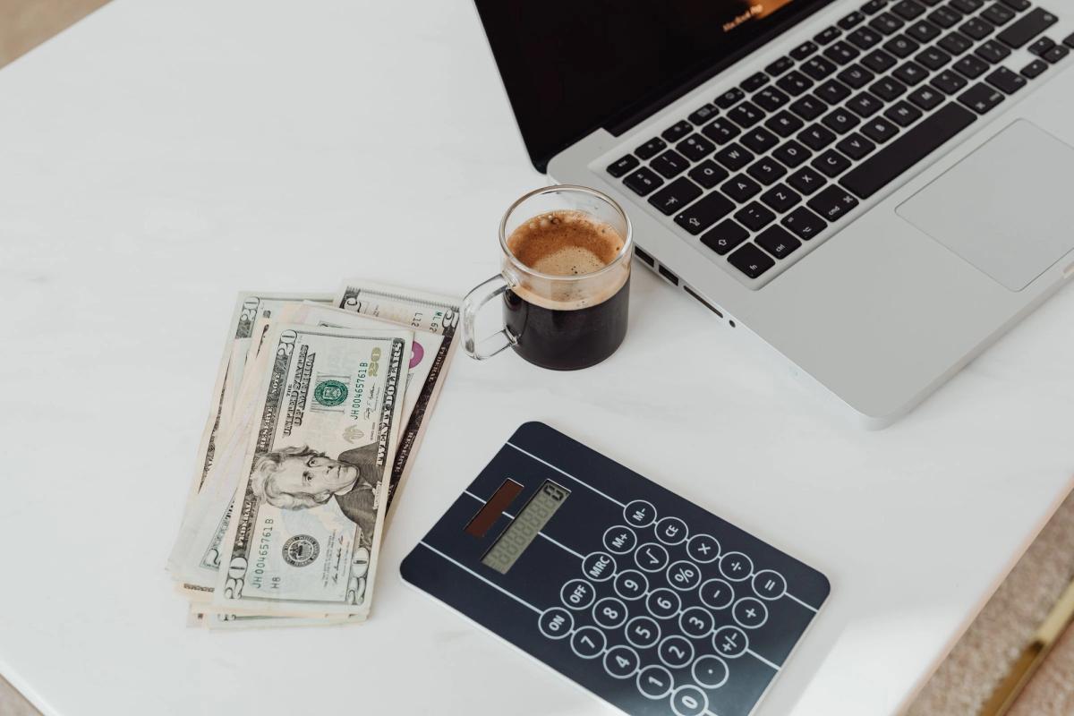 A calculator, a stack of twenty dollar bills, a glass mug of coffee, and a laptop on a white desk