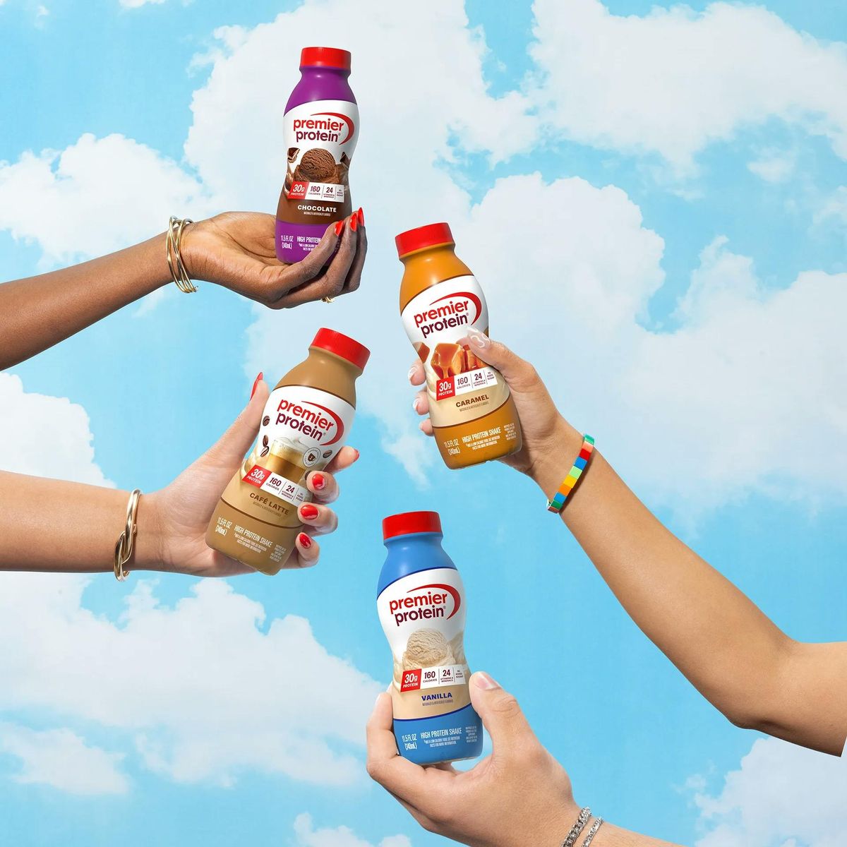 Hands holding chocolate, caramel, café latte and vanilla Premier Protein shakes against a blue sky.