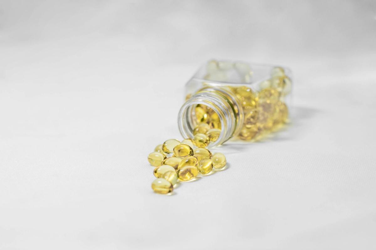Small golden softgels spilling from a clear glass jar onto a white surface.