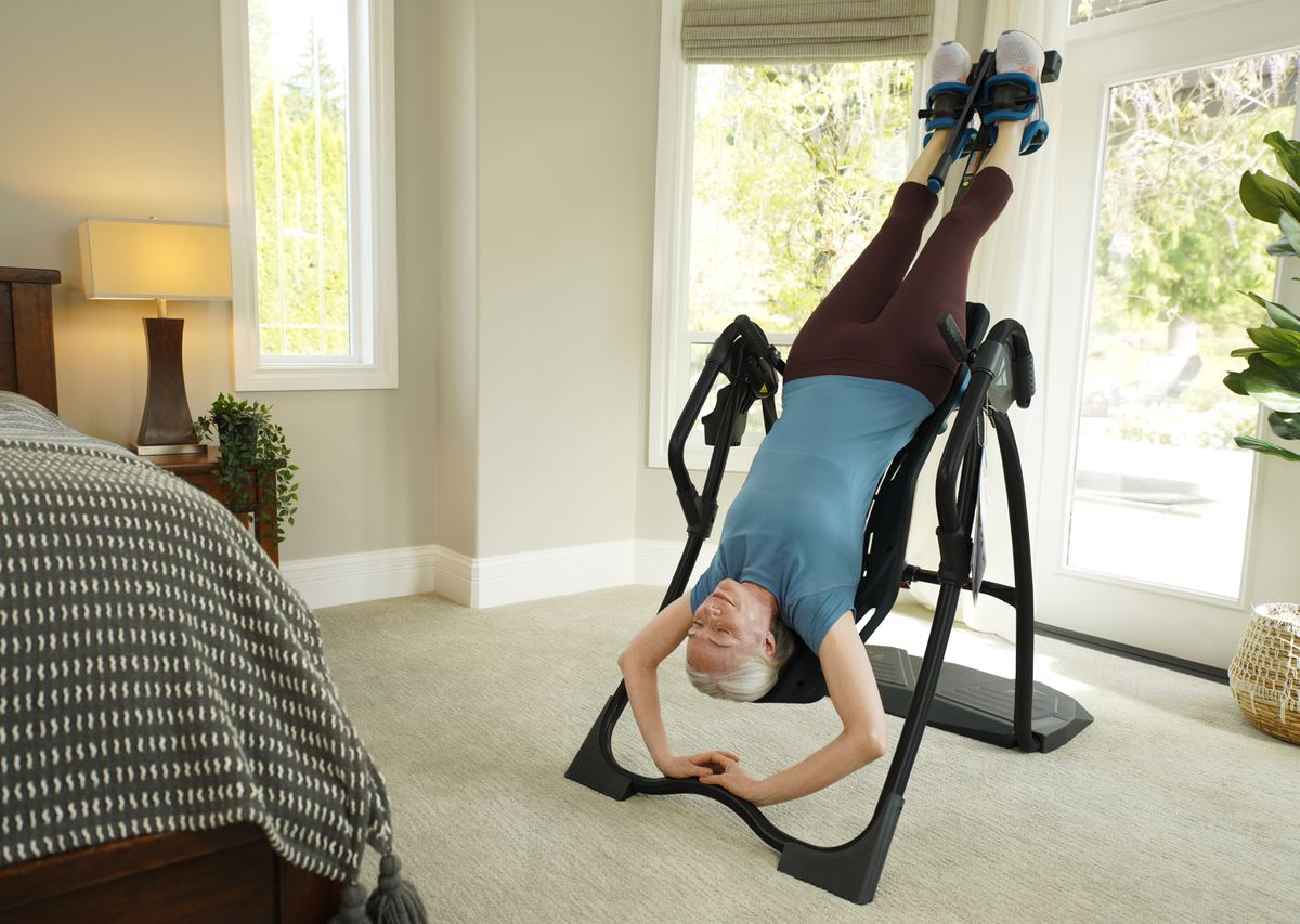 A woman hangs upside down on an inversion table in a bright bedroom