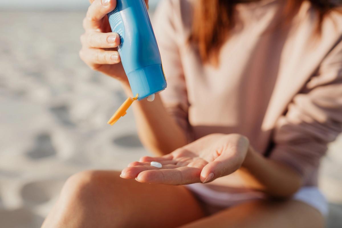 A person dispenses sunscreen lotion from a blue bottle into their palm while sitting on a sunny beach