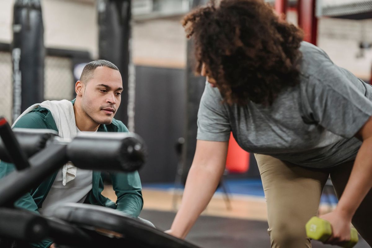 A personal trainer watches a woman lift a dumbbell in a gym.