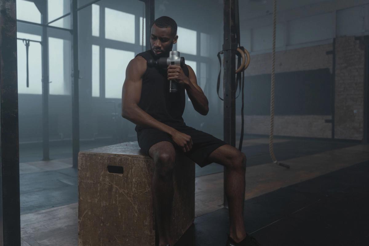 Athlete using a percussion massage gun on his shoulder in a gym