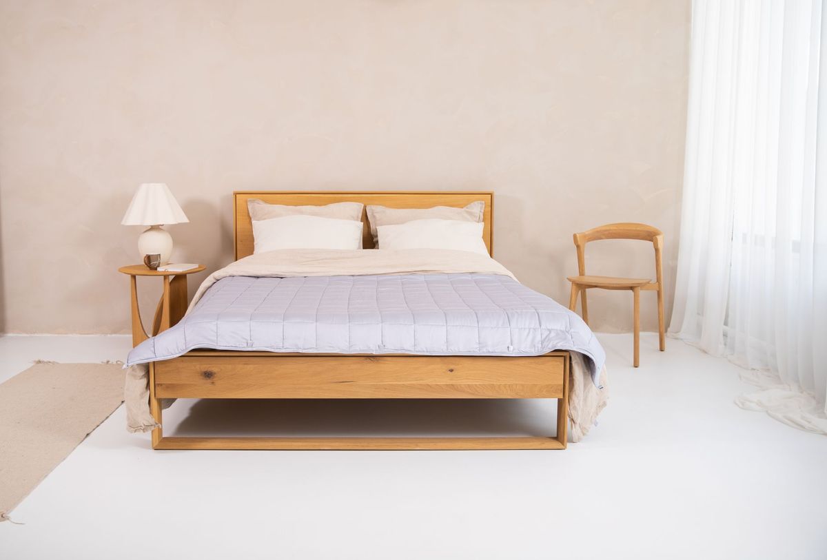 A pale gray quilted blanket spread across a wooden bed in a bright bedroom.