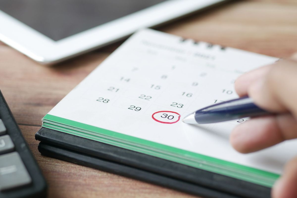 A hand circling the thirtieth on a desk calendar in red pen, with a calculator and tablet nearby.