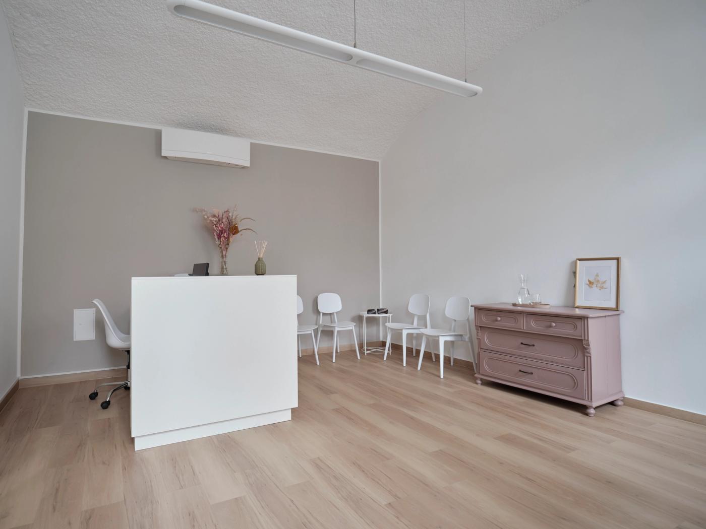 Blick von der Eingangstür in den Warteraum: links das schlichte weiße Empfangspult, rechts an der graubeigen Wand die mauve aufgearbeitete Kommode, dazwischen weiße Stühle.