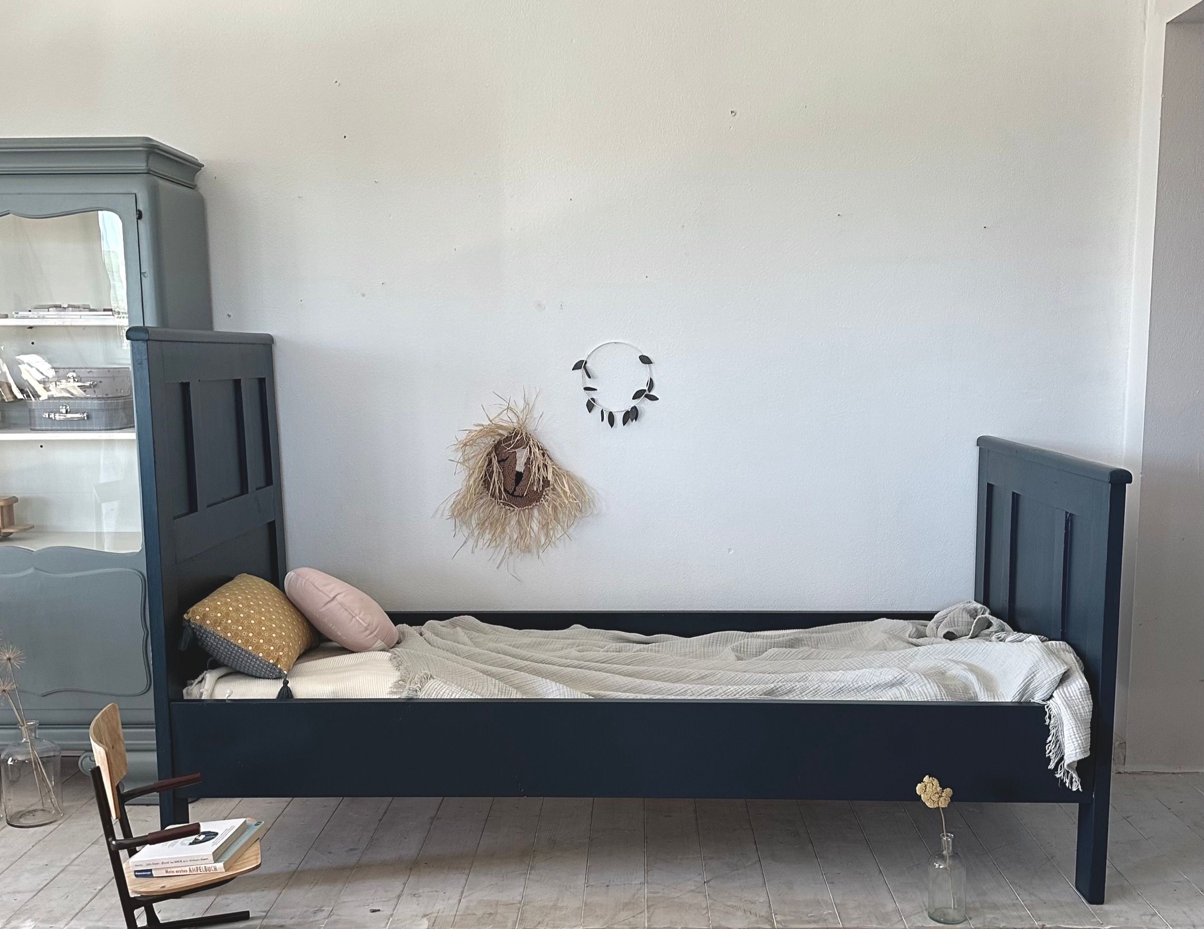 Aufgearbeitetes Vintage Kinderbett in Dunkelblau mit Pölstern und Decke, daneben eine Vitrine
