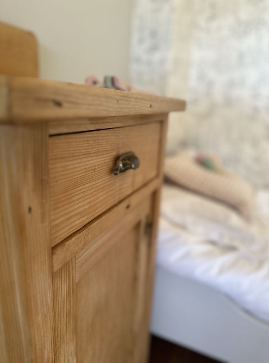Ein Nachtkästchen aus hellem Weichholz mit einer Lade und einer Tür, dahinter unscharf das frisch bezogene Kinderbett.