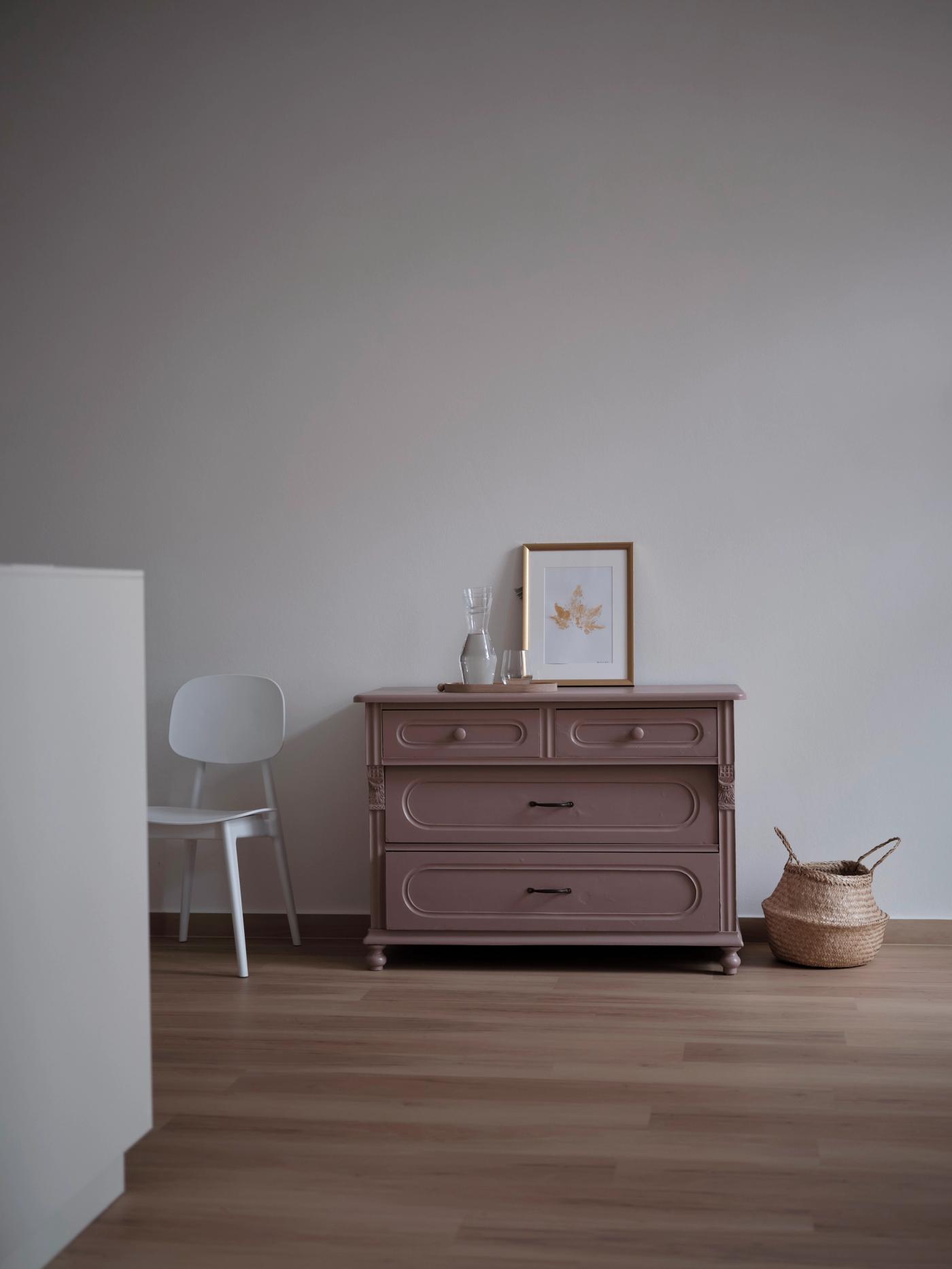 Die aufgearbeitete Kommode in gedecktem Mauve, darauf ein gerahmtes Bild und eine Glaskaraffe, daneben ein geflochtener Korb.