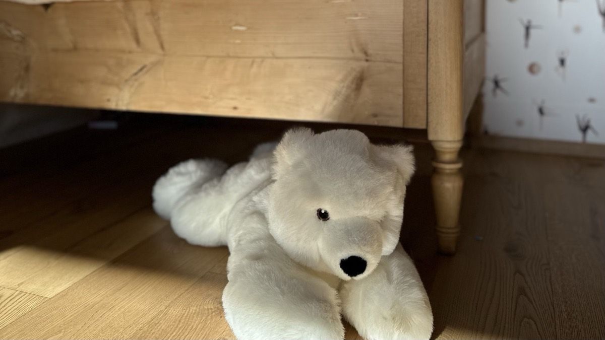Ein weißer Plüscheisbär liegt auf dem Holzboden vor einem alten Kinderbett aus hellem Holz, rechts daneben eine Tapete mit kleinen Tänzerinnen.