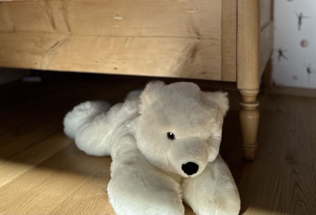 Ein weißer Plüscheisbär liegt auf dem Holzboden vor einem alten Kinderbett aus hellem Holz, rechts daneben eine Tapete mit kleinen Tänzerinnen.
