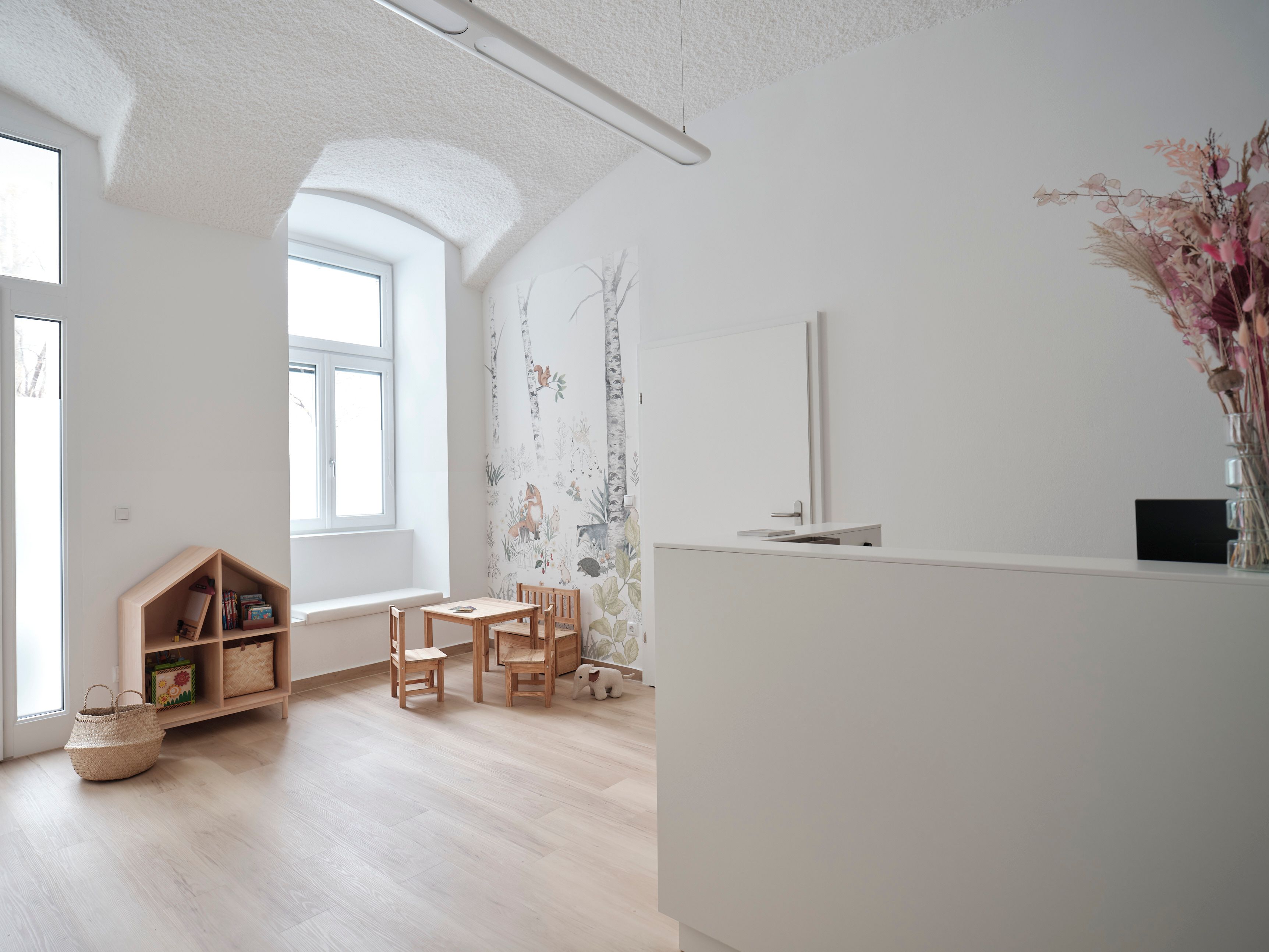 Der Warteraum der Kinderarztpraxis unter einer weißen Gewölbedecke: links ein Regal in Hausform und ein Kindertisch vor der Waldtapete, rechts der weiße Empfangstresen.