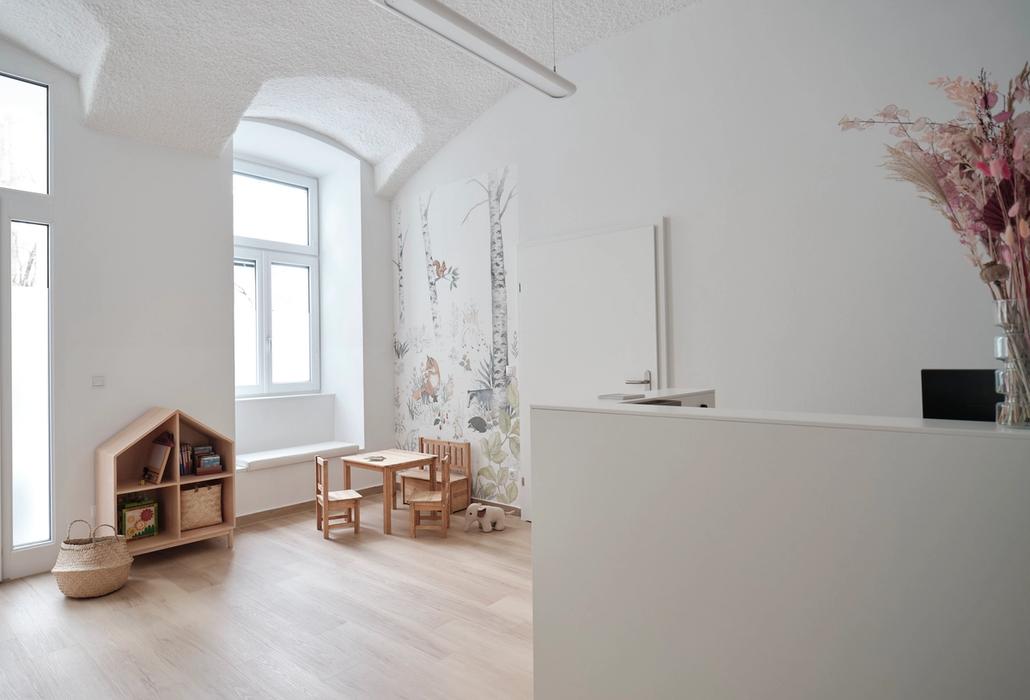 Der Warteraum der Kinderarztpraxis unter einer weißen Gewölbedecke: links ein Regal in Hausform und ein Kindertisch vor der Waldtapete, rechts der weiße Empfangstresen.