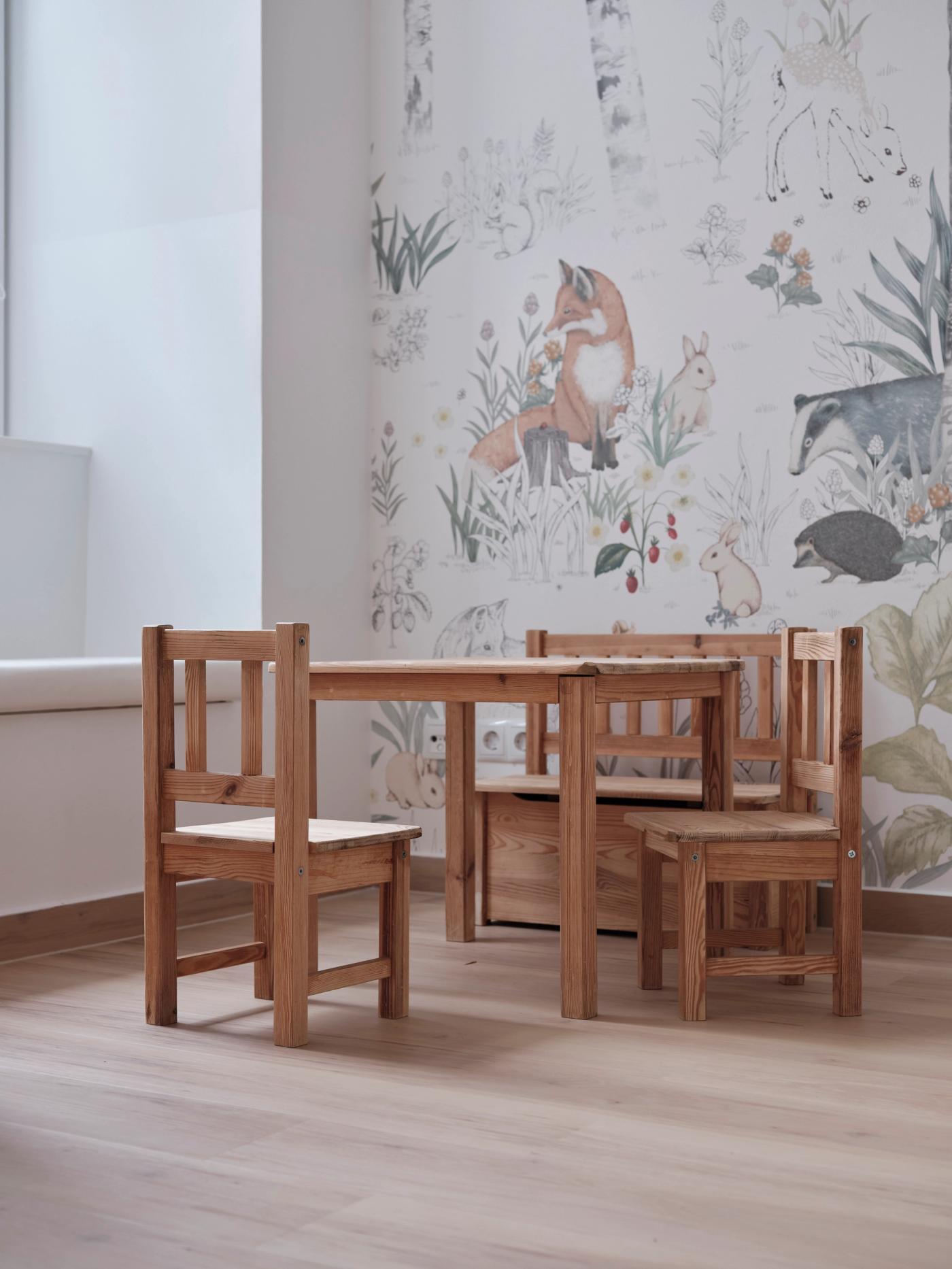 Ein kleiner Tisch mit drei Kinderstühlen aus Naturholz vor einer Tapete mit Waldtieren.