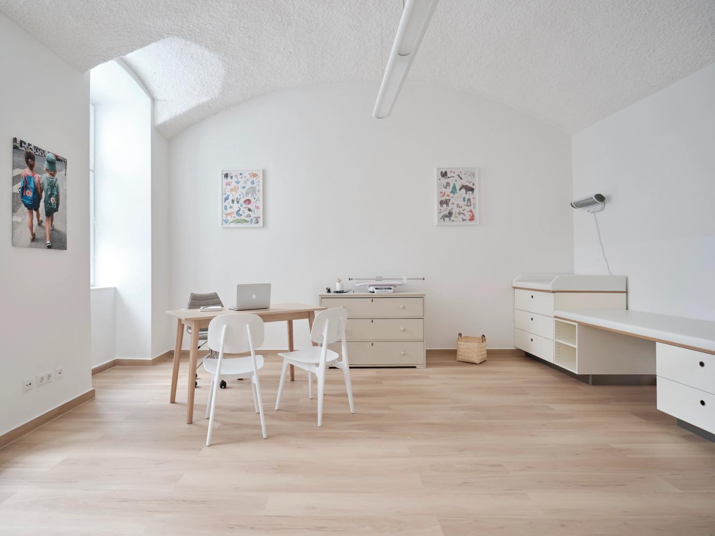 Der Behandlungsraum: ein heller Holztisch mit zwei weißen Stühlen, dahinter eine Kommode und die Untersuchungsliege, an den Wänden gerahmte Bilder.