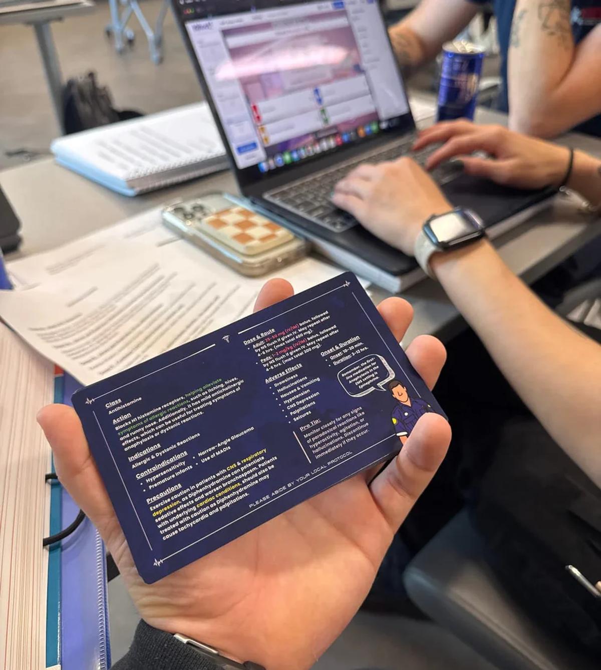 A student holds a Paramedic Flash pharmacology card beside a laptop