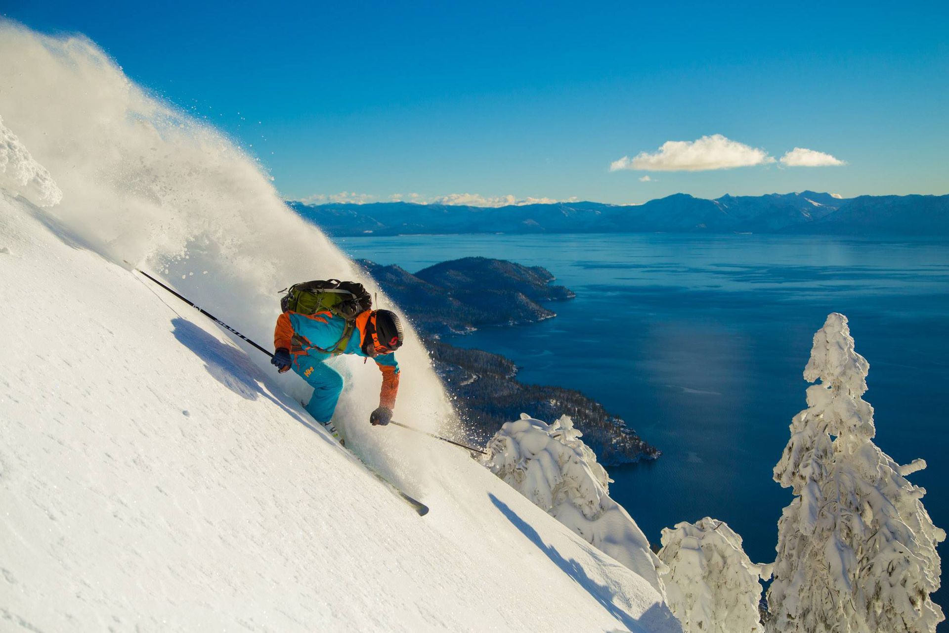 Skier in an orange and teal jacket throwing powder spray on a steep slope high above Lake Tahoe