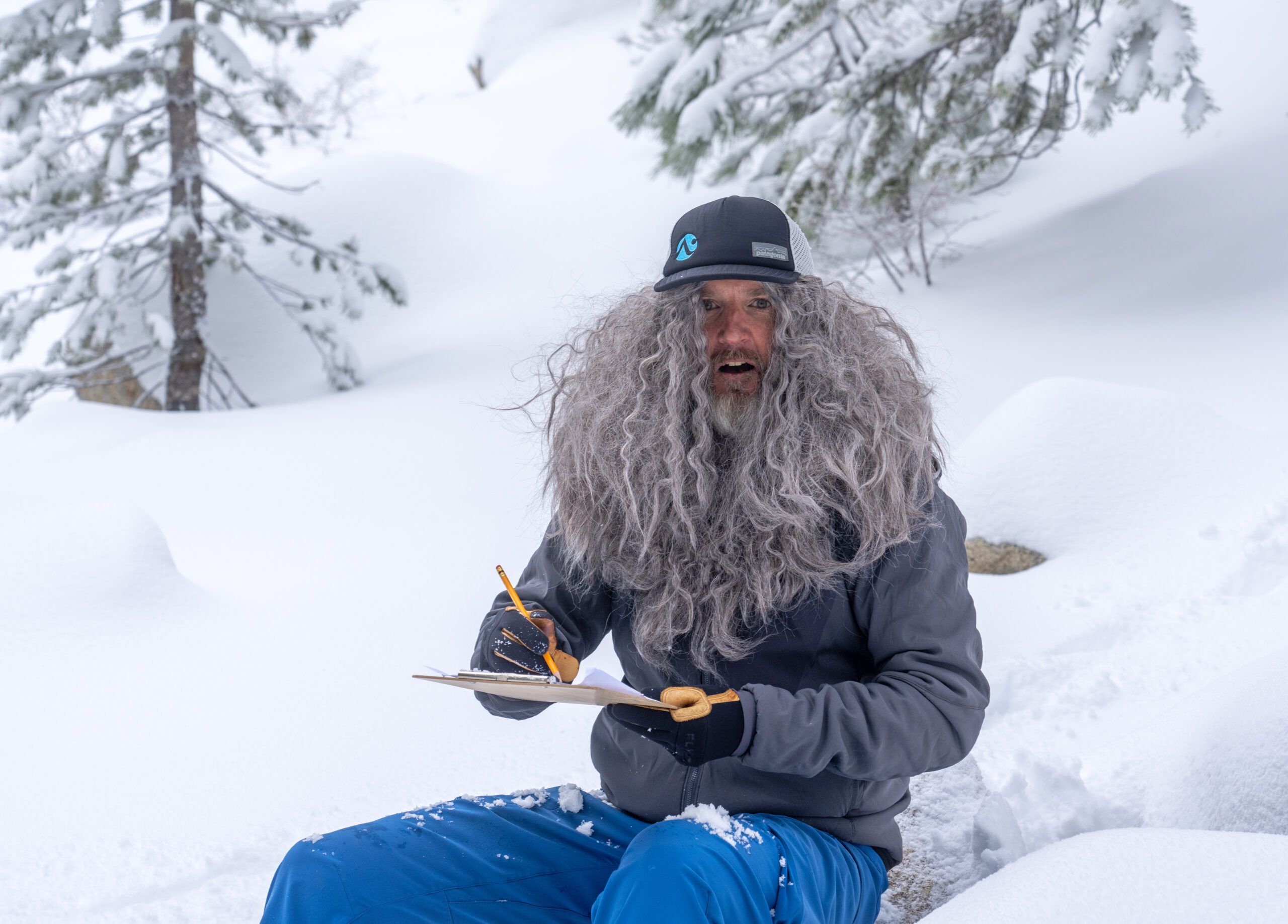 Backcountry skier in a gray wig and beanie sitting in the snow writing on a clipboard