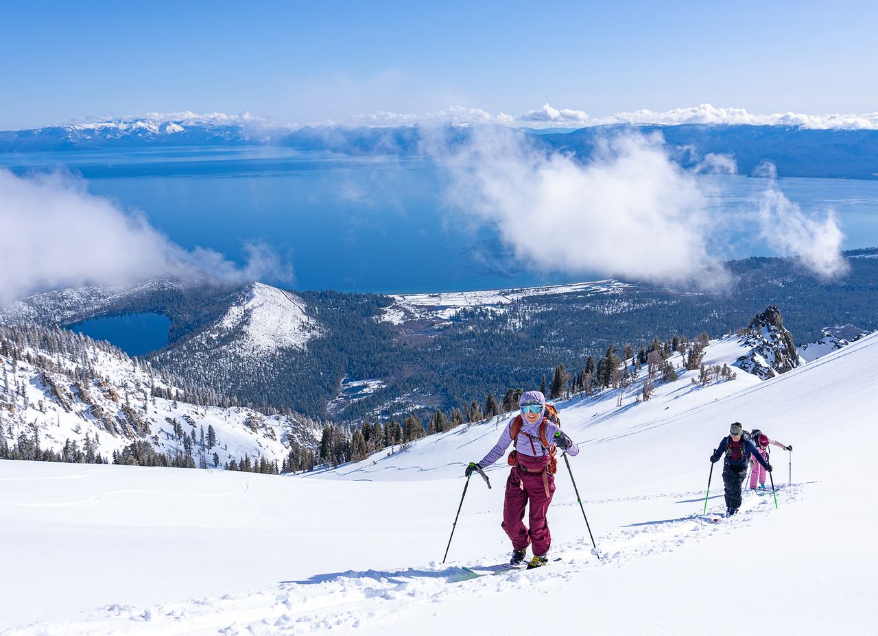Ski tourers skinning up an open snowfield with Lake Tahoe and clouds behind them