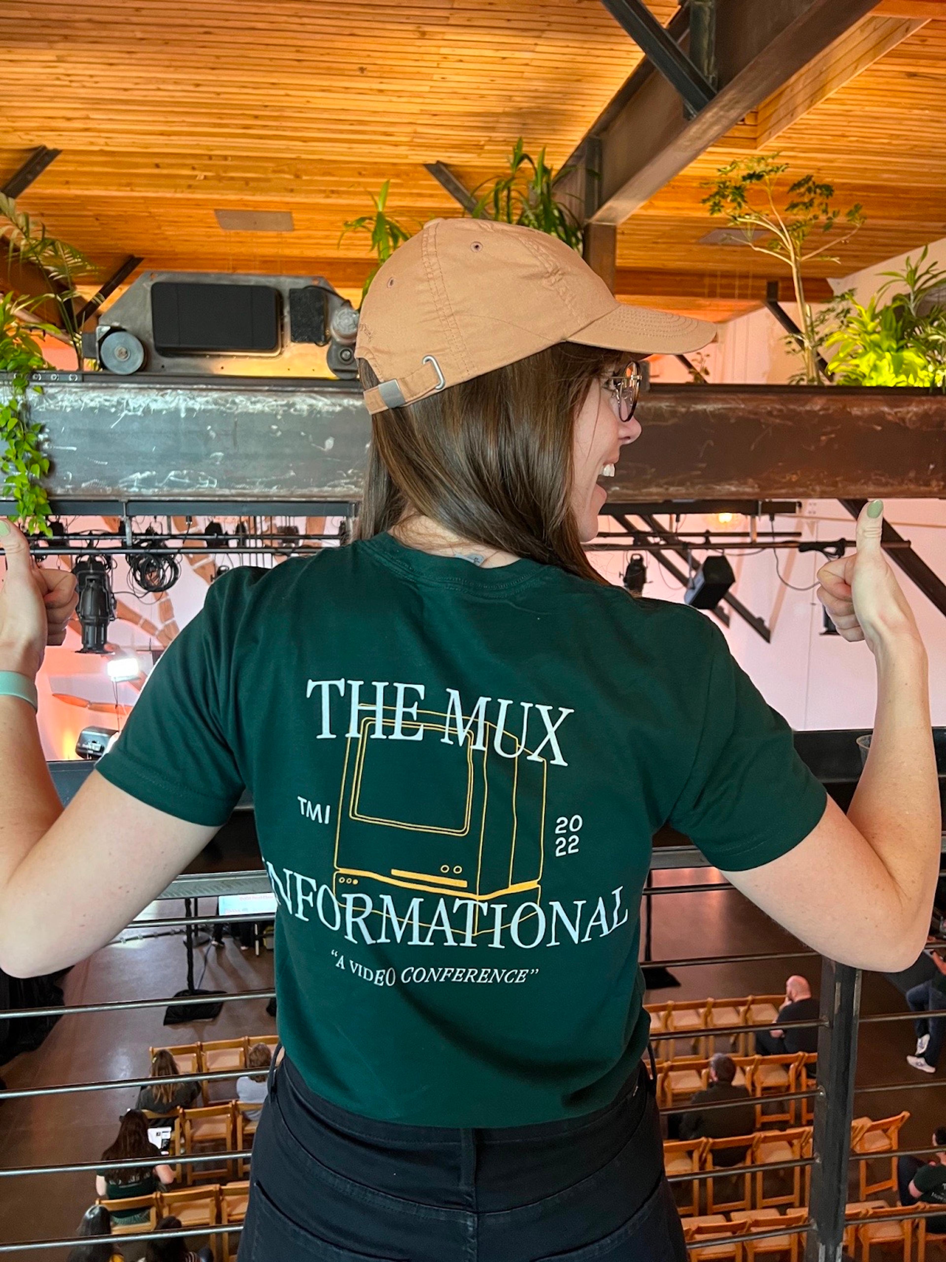 A woman wearing a dark green t-shirt with the words, "The Mux Informational. TMI 2022. A video conference." on it. She is facing away from the camera, with two thumbs up, looking behind her excitedly. She also wears a brown hat and black jeans.