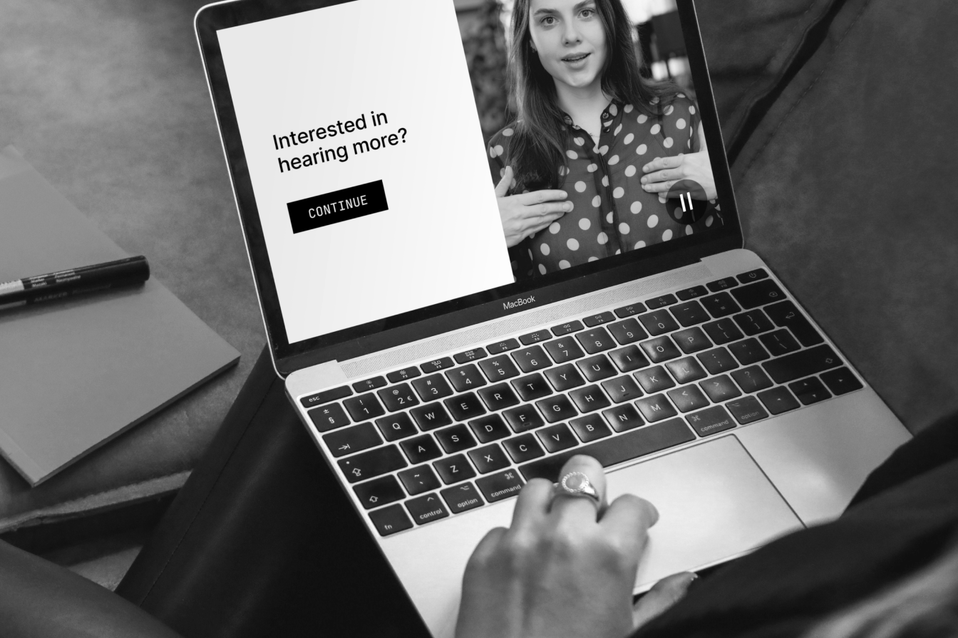 Black-and-white photo of a laptop playing a video of a woman, with an on-screen prompt asking Interested in hearing more? and a Continue button